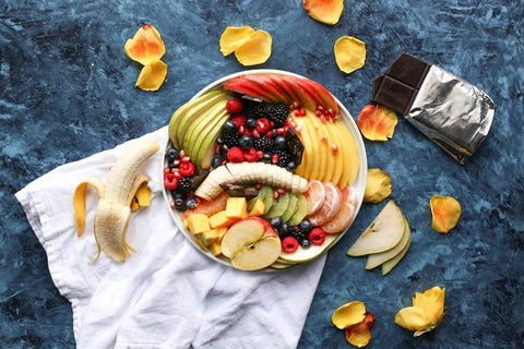 Bowl of assorted fresh fruits including banana, apple, berries, kiwi, and mango with a partially peeled banana and chocolate bar on a blue textured background for optimal training nutrition.