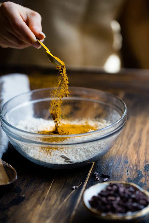 Hand sprinkling spice over flour in a glass bowl for healthy sports nutrition recipe focused on fats.